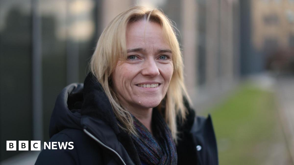 A woman with blond hair, wearing a dark jacket and smiling into the camera while standing outside an out of focus building