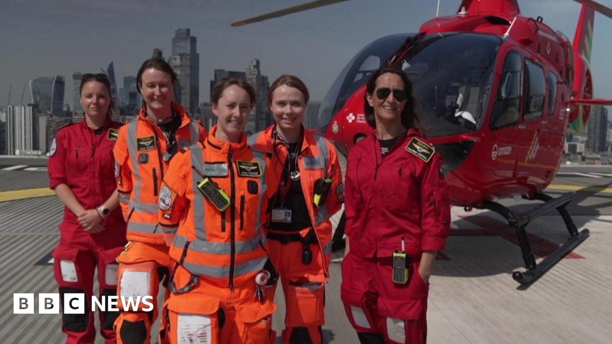 Group photo of the five female crew members on top of a building with the red helicopter behind them and a London skyline in the background