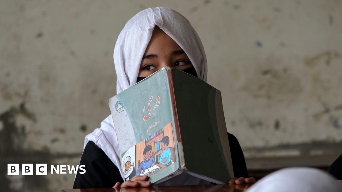 A girl with her hair covered with a white scarf hides her faces behind a reading book with a picture of two young boys on it. She is sitting with her back to a concrete wall in what appears to be a classroom