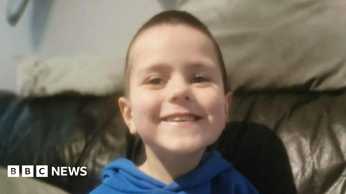 A young boy smiling at the camera. He is wearing a blue hoodie and sitting on a leather sofa.