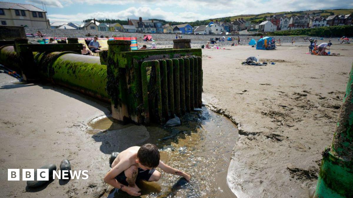 A boy plays in a stream in front of a discharge pipe on a sunny beach in Wales, with sand and sunbathers in the background. 