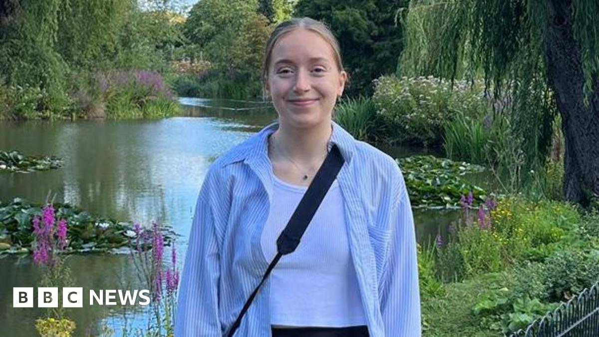Libby smiling into the camera in front of a pond with lily pads, plants and flowers. She has blonde hair which is tied back and is wearing a blue and white striped shirt with a white tank top underneath. She is also wearing a black cross-body bag and a...