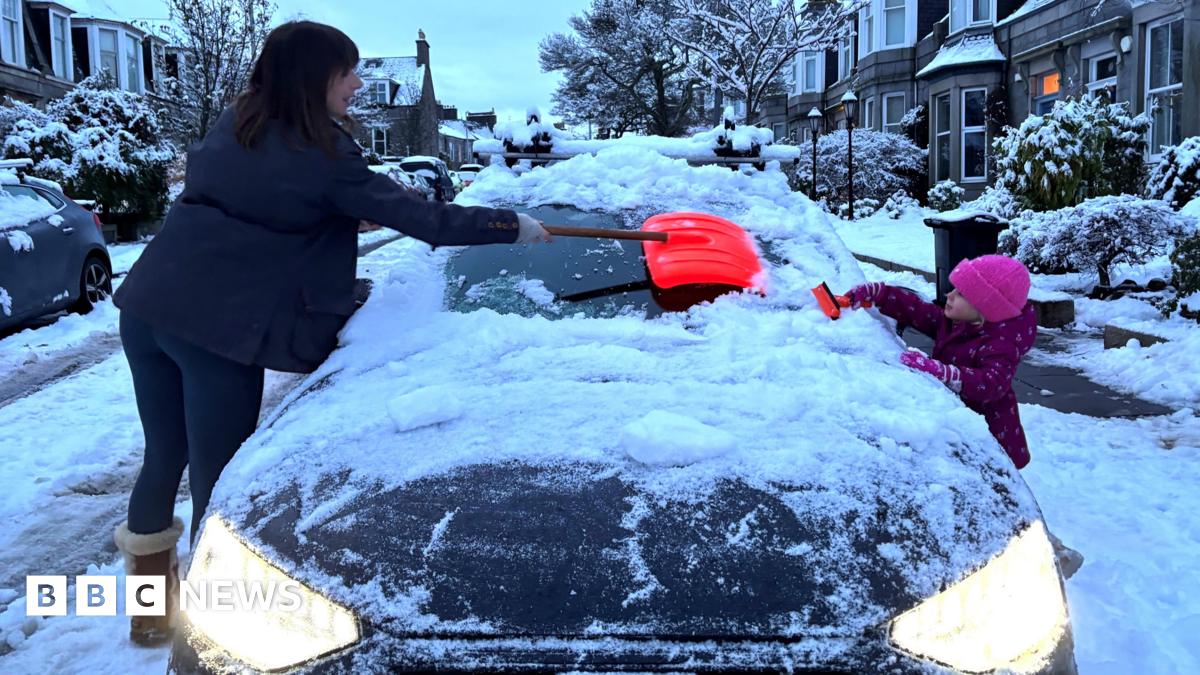 A mother and daughter clear the snow-laden windscreen of a car in the early hours. The car is on an upmarket residential street with sandstone properties, trees and bushes.