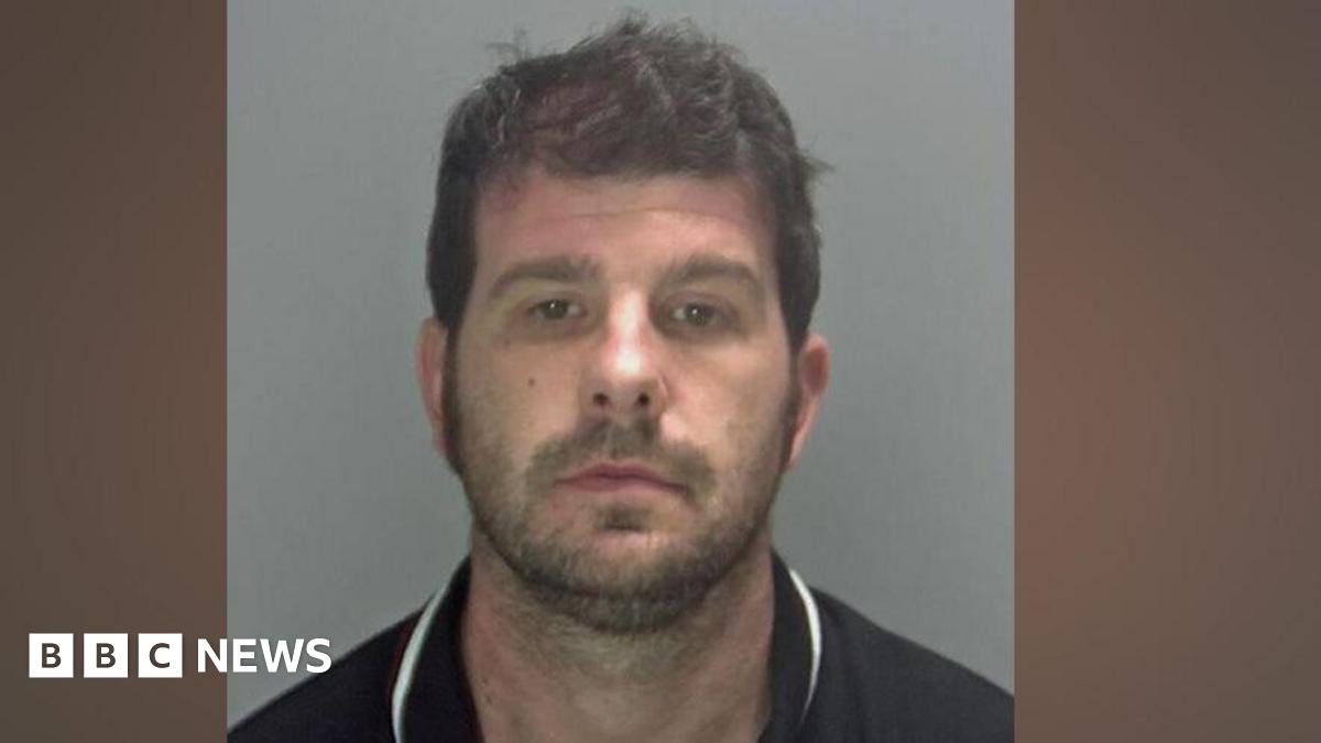 Stuart Worby in police custody, wearing a black or navy blue polo shirt with a white trim on the collar. He has black hair and stubble.