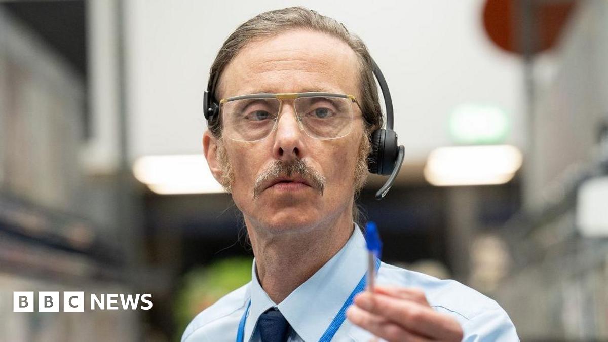 Mackenzie Crook as a shop worker with headset and glasses. he wears a shirt and tie and points with a pen.
