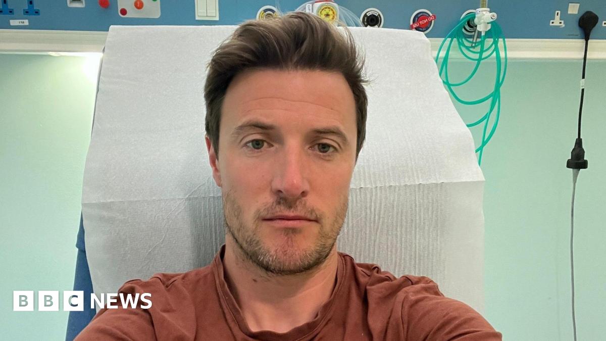 A man in a brown T-shirt takes a selfie in a hospital bed. He is not smiling.