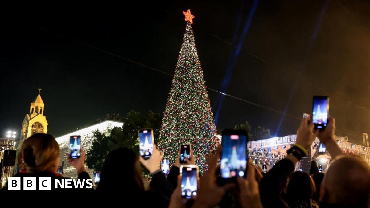 A giant, lit up Christmas tree with people holding phones standing around it 