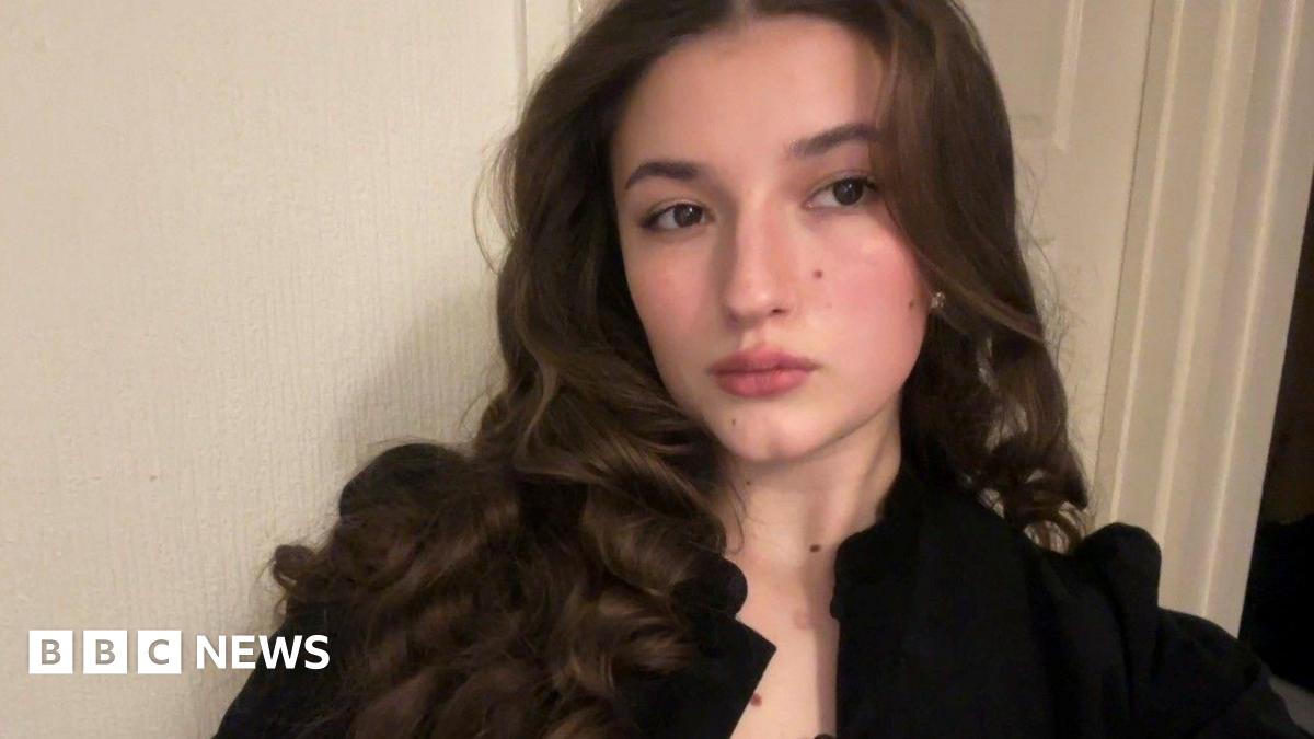 A brunette girl with curly hair, looking towards the camera with a straight expression. She wears a black open blouse and stands against a plain white back drop.
