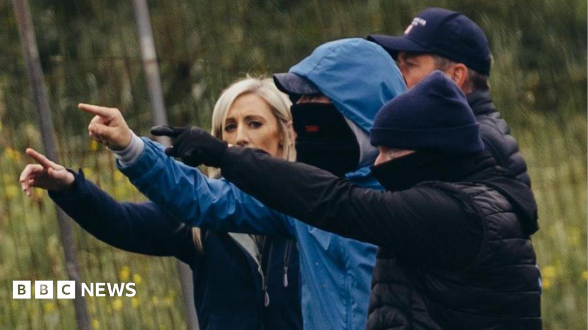 Carla Lockhart standing beside four men. They are all pointing their fingers with their arms in the air. 