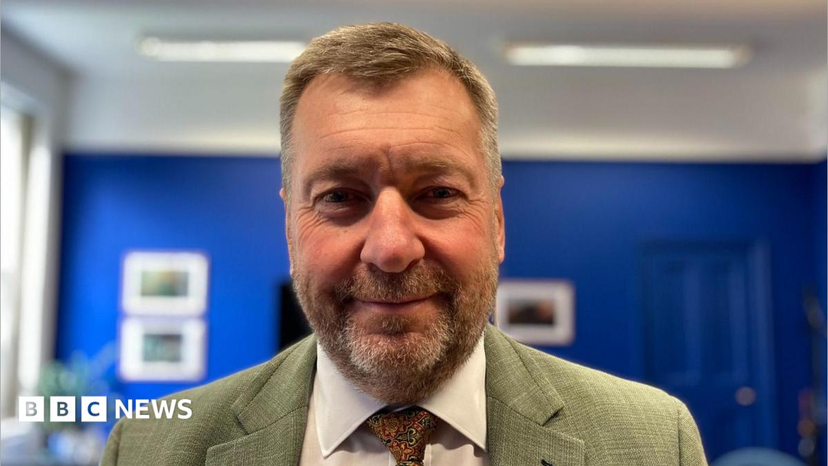 A head and shoulders photograph of Lincolnshire County Council leader Sean Matthews standing in a room with blue walls and a white ceiling.  He is wearing a green blazer with a white shirt and paisley tie.