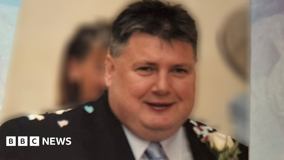 John Smyth, a well-built man with short, dark hair, wearing a black suit with white shirt and blue tie. He appears to have confetti scattered on his shoulders and a white flower in the lapel of of his jacket.