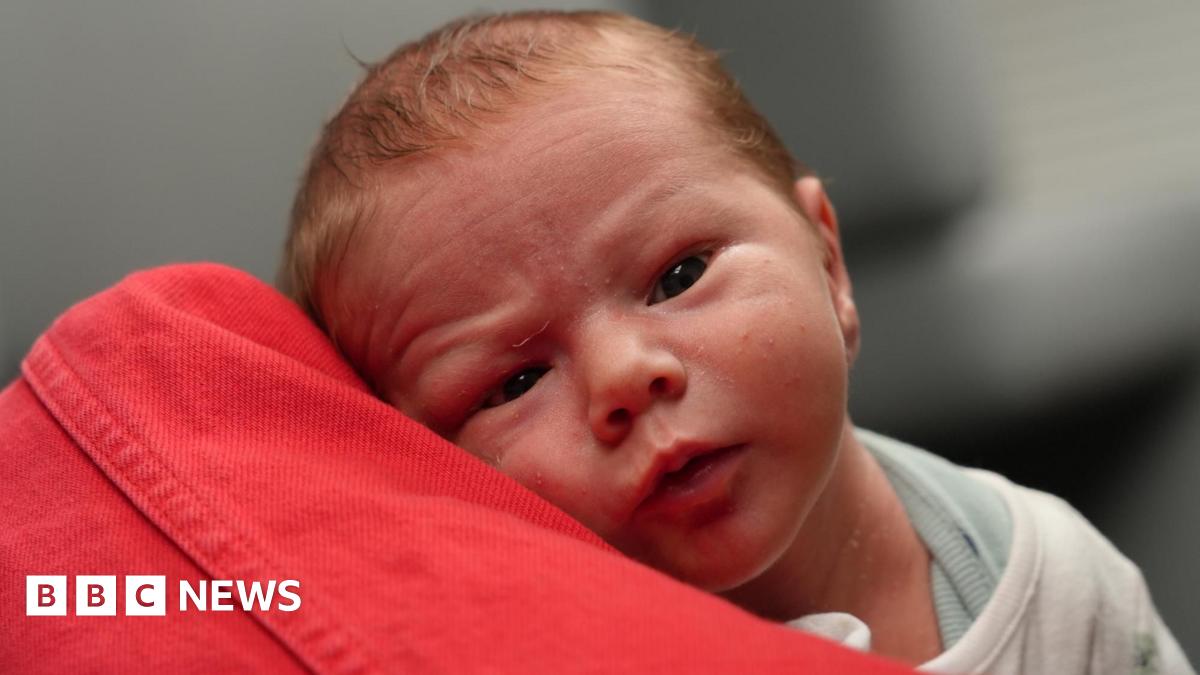 Baby Henry wide awake looking directly at the camera. He is resting on the arm of his grandmother who is wearing a red denim jacket. You can only see her arm. Henry is wearing a vest and a cream bodysuit. He has a tiny wrinkled forehead and is looking...