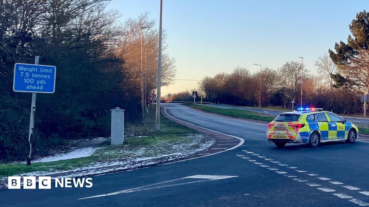 The A120 road which runs through Essex. On the carriageway there is a parked yellow and blue fluorescent marked police car which is blocking the way for any traffic to get down the road. On the right of the picture is a green sign pointing people to St...