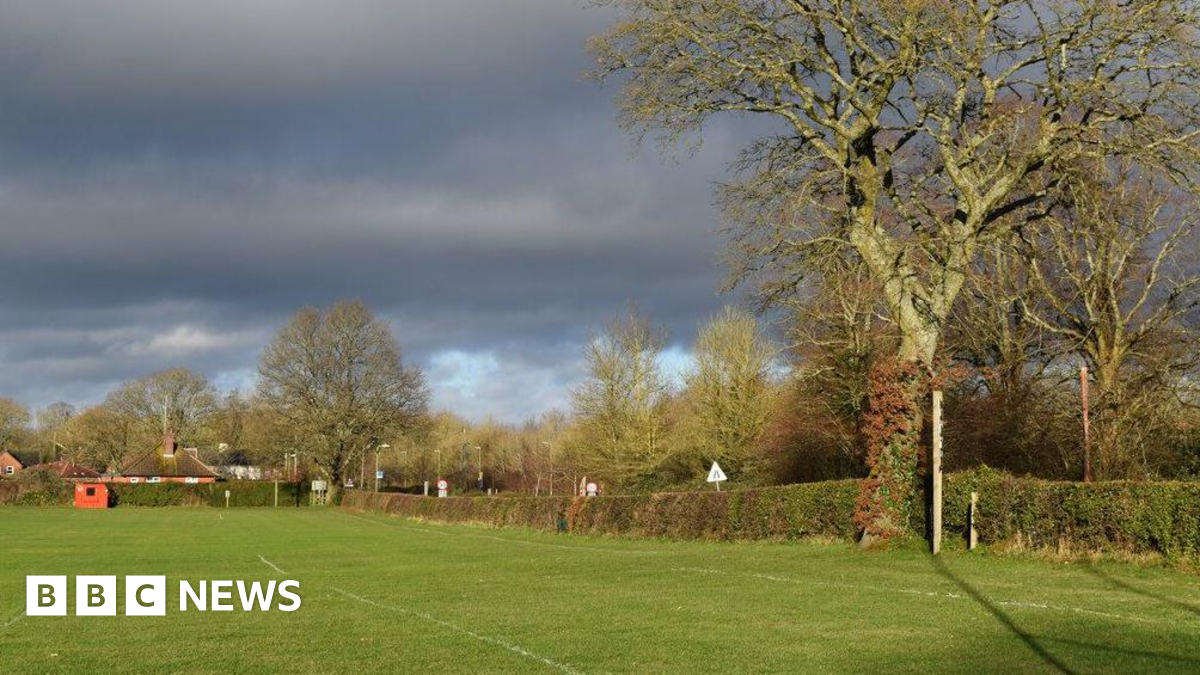 View along the edge of the recreation ground. A playing field is in the foreground, with trees and a hedge to the right.
