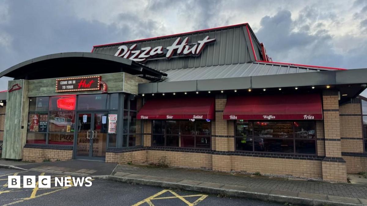 The exterior of a Pizza Hut restaurant with a grey roof displaying a large 'Pizza Hut sign' and maroon window awnings on the windows.