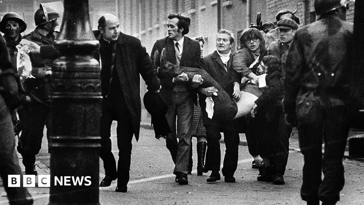 A picture taken on 30 January 1972 - Bloody Sunday. A group of men are carrying a lifeless body - they are accompanied by a priest waving a wife handkerchief. There are soldiers at the side of the picture and in the foreground.