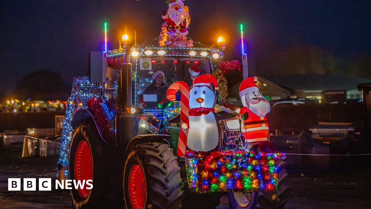 A tractor decorated in festive colourful lights at night time. It has a blow up Santa Claus and snowman attached to its front. 