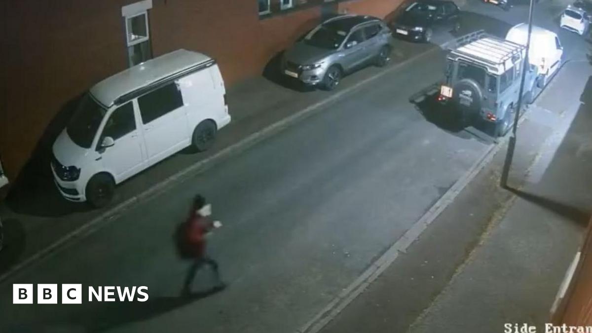 Blurry CCTV footage of a slim man who is dressed in a maroon jacket, black beanie hat and dark trousers. He is walking along a deserted street at night.