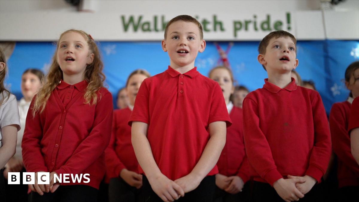 Students at Mersey Drive Community Primary School sing in their school hall. It's a close-up shot of three singing primary age children, two boys centre and right, and one girl on the left, all in red school uniform tops. More children stand behind the...