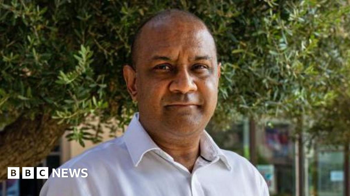 Sanjay Shah standing outside underneath trees in what appears to be a garden in a built up office area - he is a middle aged man, with a white shirt and thinning hair. He is looking just past the camera with a neutral expression. 