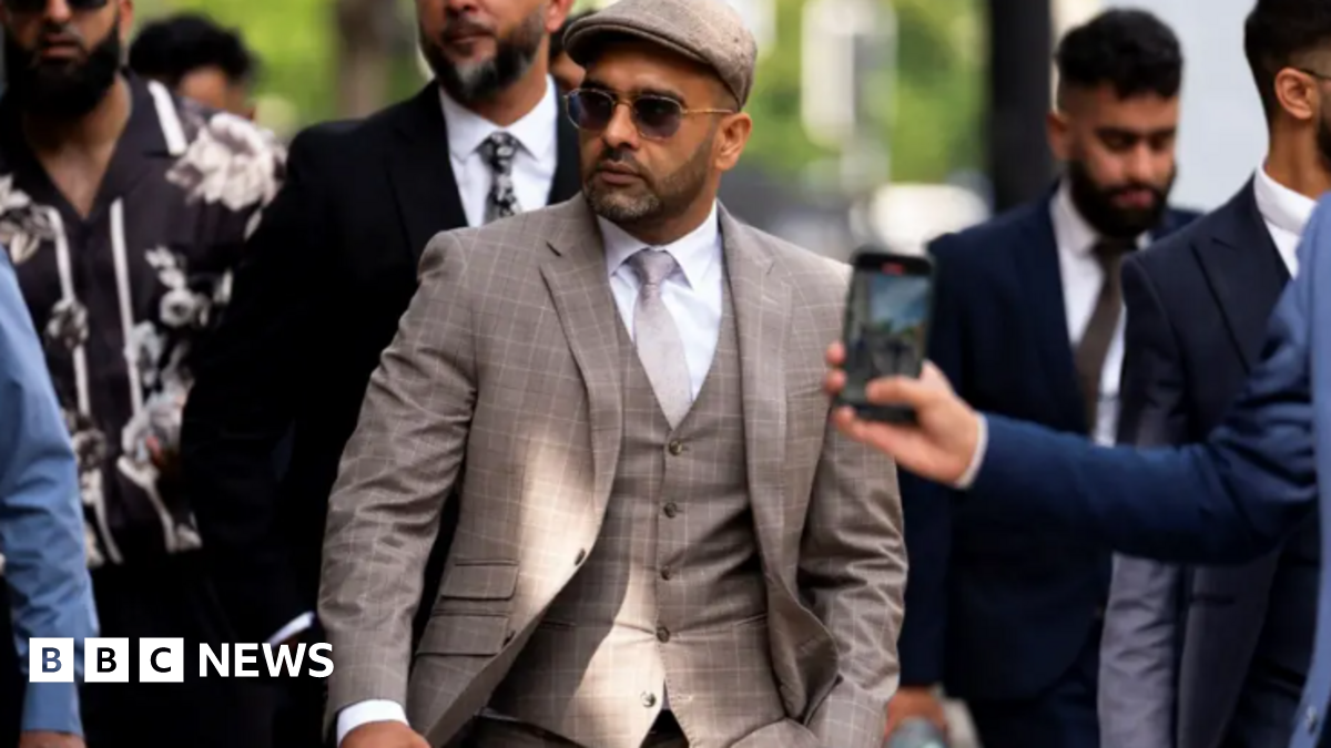 Akhmed is wearing a beige patterned suit with a waistcoat, white shirt, flat cap and sunglasses. He is walking on a street and has several people flanking him.