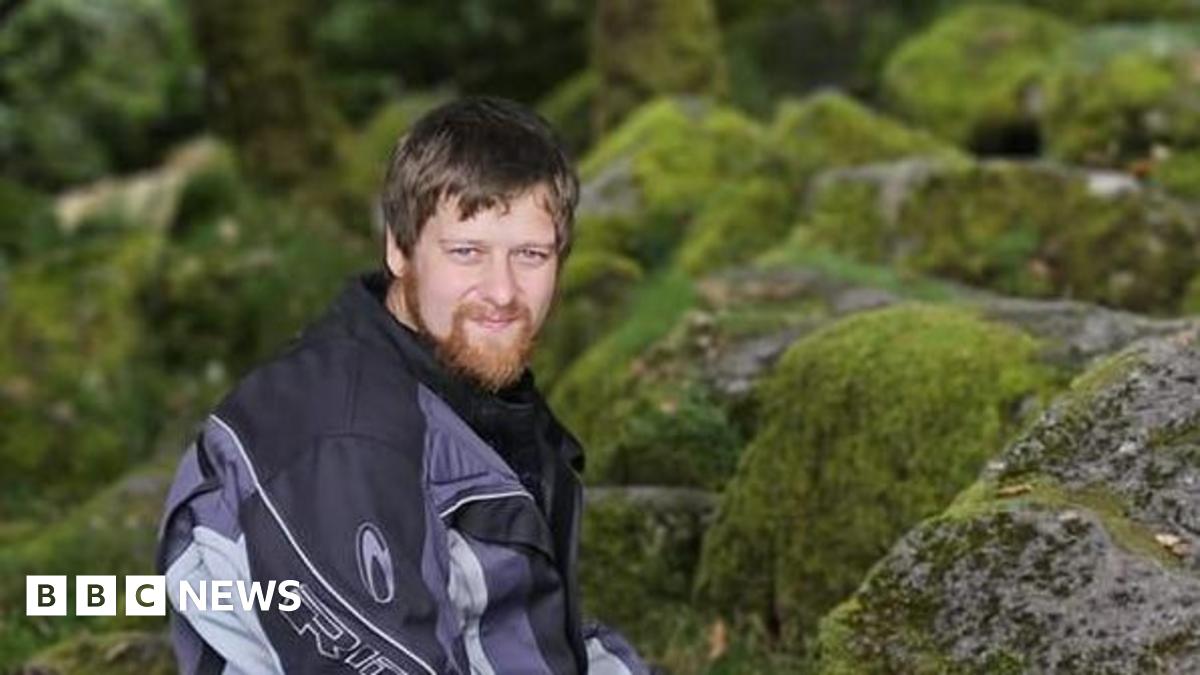 Ben Rowe, with a beard, is wearing a motorcycling jacket and sitting on rocks covered in moss in his former Facebook profile picture