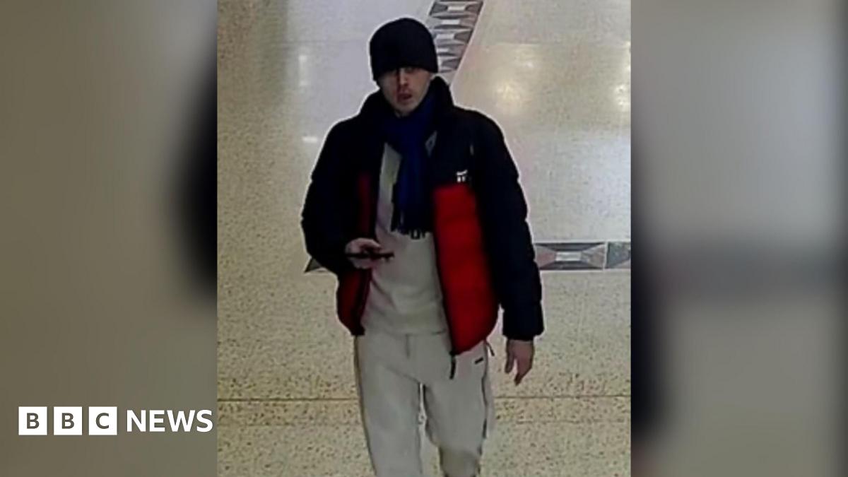 A CCTV image of a man in a grey tracksuit, black and red coat, dark scarf and black beanie. He is looking up and holding a phone.