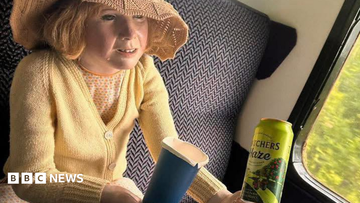 A child mannequin with a straw hat and cardigan sitting on a train. The mannequin is holding a can. The train appears to be moving.  