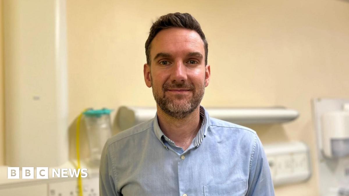 Dr Mike Blaber in a blue shirt in a hospital ward with equipment in the background