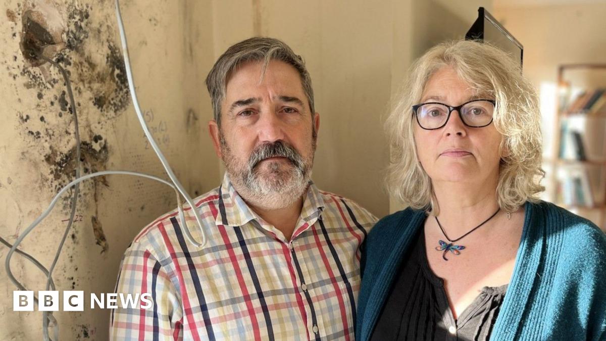 Tony and Bec Wadley standing next to a wall covered in black mould with electrical wires and sockets hanging from it