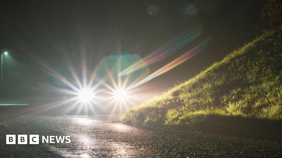A generic picture showing a car's headlights glowing at night on an empty country road, with a grassy bank on the right.
