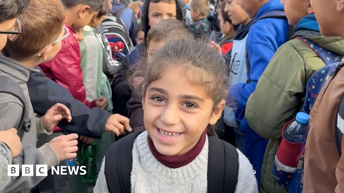 A child with her hair tied back smiles, with backpack straps and a cream jumper. Other children surround her.