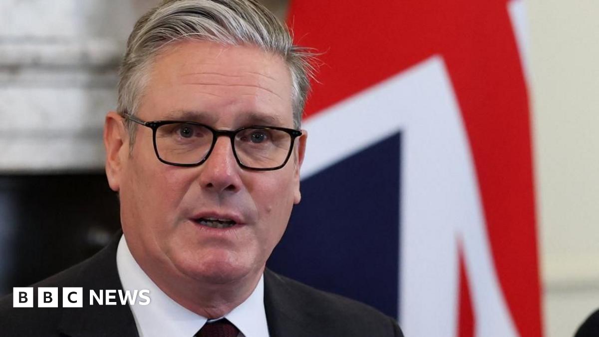 Keir Starmer is pictured in front of union jack, wearing a black suit with a dark red tie and glasses with his mouth slightly open, his face stern.