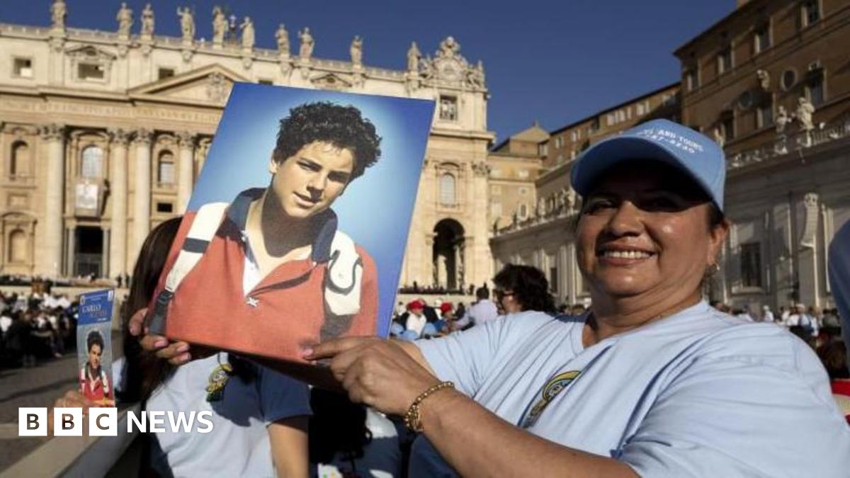A woman in a crowd at the Vatican, wearing a white T-shirt and a pale blue baseball cap holds up a portrait picture of curly-haired Carlo Acutis, wearing a red jumper with blue collar, with the white handles of a backpack showing