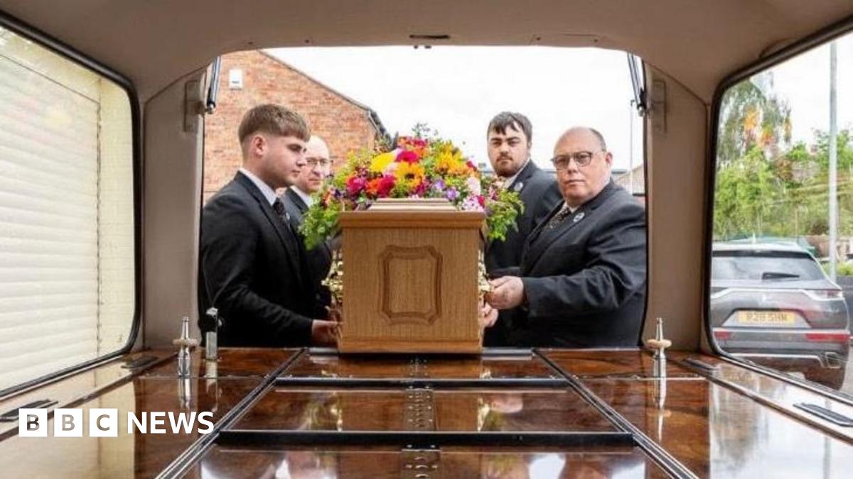 A funeral director team putting a coffin with flowers laid on top into a hearse. They are in a residential area and are all wearing suits. The view is from inside the hearse