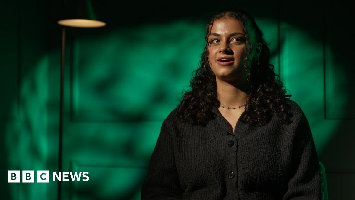 Ruby Greensides sits facing the camera in a studio interview, wearing a dark cardigan, with green light patterns behind her.