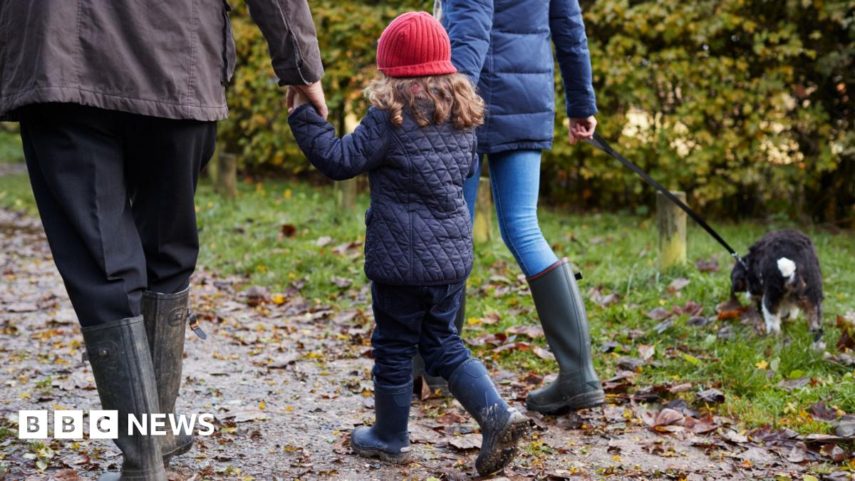 A child wearing a blue coat and red hat with face turned away from the camera walks between two adults