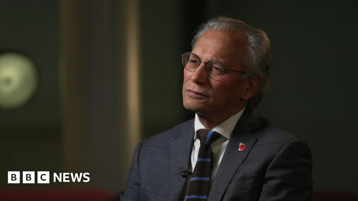 Samir Shah wears a grey blazer, the lapel studded with a Remembrance poppy, a white shirt and a brown and blue stripped knitted tie.  