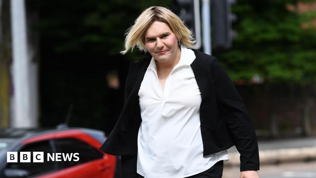 A picture of a transgender woman with short straight blonde hair, wearing make up, hoop earrings, a white blouse and a black cardigan. Katie Wallis is walking into court and looking at the camera.