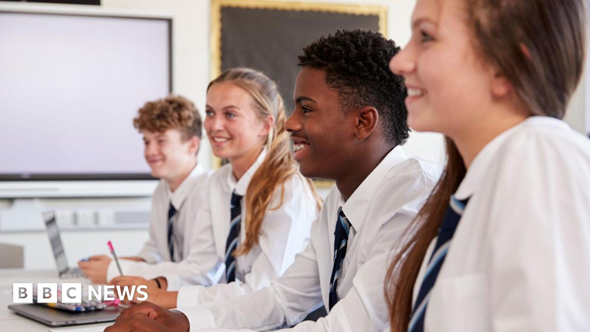 Four secondary school students sit at a row of desks. They are all smiling towards something that is out of vision. All four students wear the same uniform - a white long-sleeved shirt with a navy, blue and white striped tie. The boy on the far-left, w...
