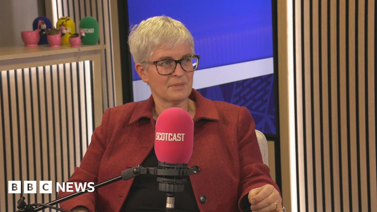 Naomi Cunningham sitting in a BBC podcast studio. She is behind a microphone with a red cover that says Scotcast. She has short, grey hair and glasses. She is wearing a red jacket