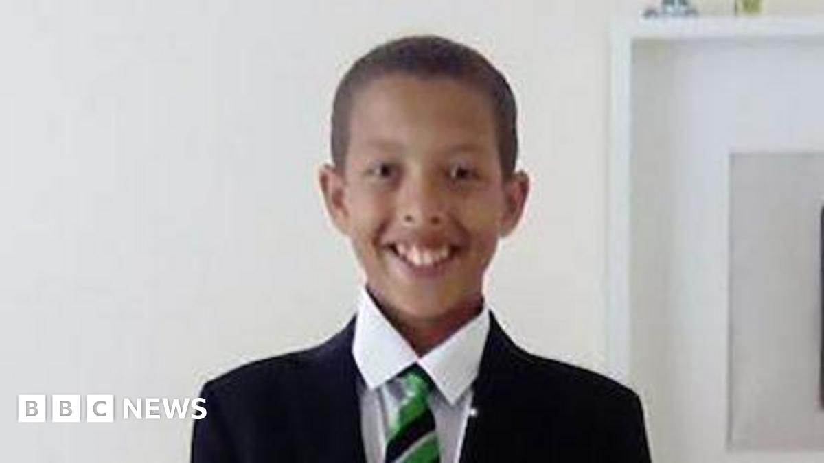 Noah smiling at the camera as he stands against a white wall. He is wearing his school uniform which is a black blazer, green and black striped tie and a white shirt. He has short black hair.