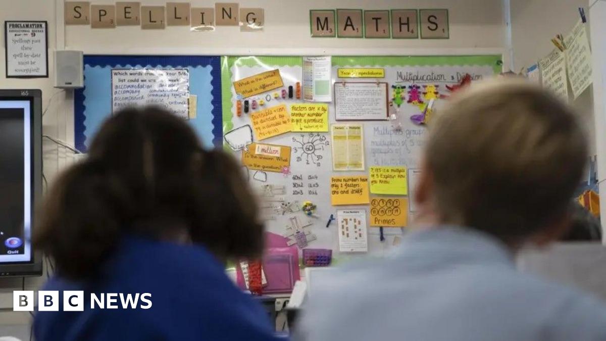 Classroom wall display with ‘Spelling’ and ‘Maths’ labels, covered in student work, notes, and diagrams, with two students visible out of focus in the foreground
