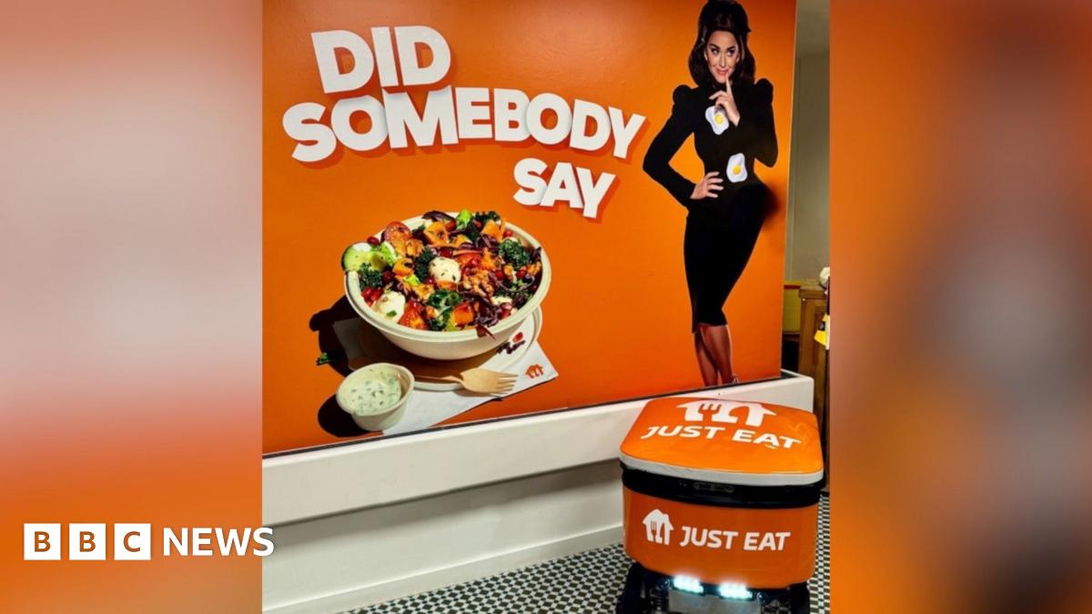 A photo of a Just Eat poster and a food delivery robot. The poster has an orange background with text that says "Did Somebody Say". Also on the poster there is a woman in a black dress with fried eggs on her dress. There is also a bowl for food with a...