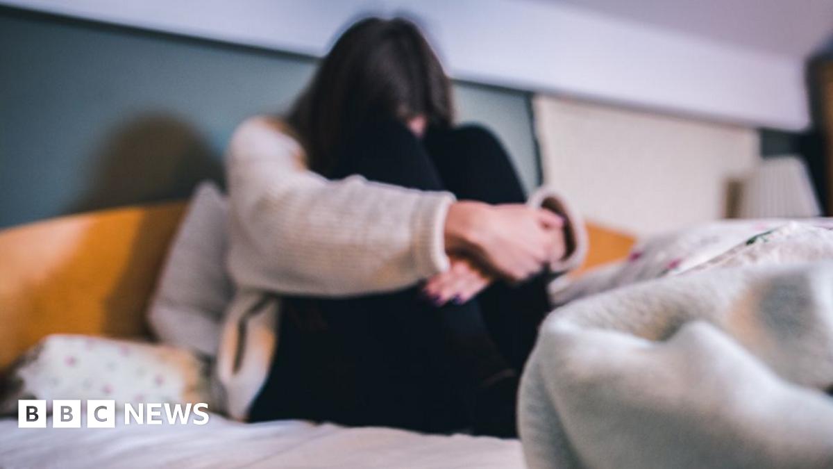 A female figure is sitting on a bed with their arms wrapped around their legs, their face cannot be seen