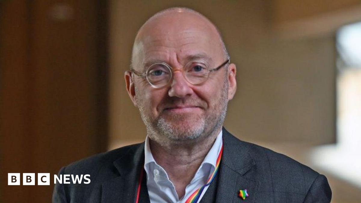 Patrick Harvie is smiling at the camera. He has a bald head and round glasses. He is wearing a grey suit, white shirt, a rainbow-coloured lanyard and a rainbow-coloured badge.