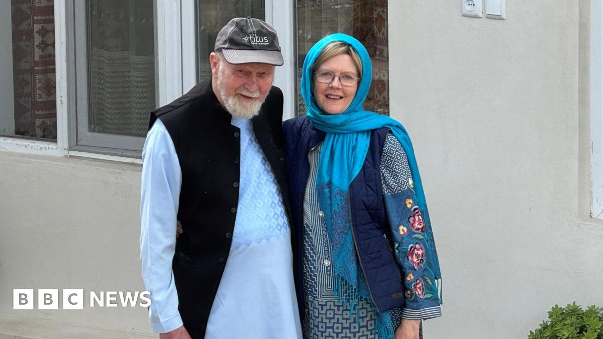 Peter and Barbie Reynolds stand together and smile at the camera. Peter Reynolds is wearing a grey cap and has a white beard. Barbie Reynolds is wearing glasses and a turquoise headscarf.