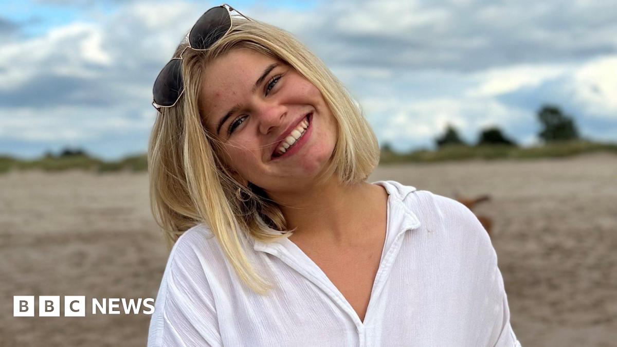 Phoenix Spencer-Horn smiling directly at the camera. She has her head tilted to the right and has blonde hair, which drapes over her shoulder and sunglasses on top of her head. She is wearing a white shirt and is standing in front of a beach, trees and...