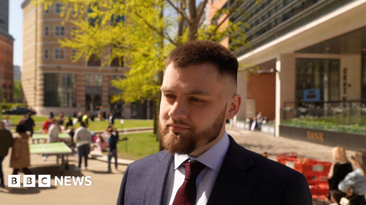 A man stands in a city square. He is wearing a navy jacket, grey shirt and maroon tie. He has spiky brown hair and manicured beard. He is looking to the right of the camera at something. Passers-by can be seen in the square behind him, the Bank pub,