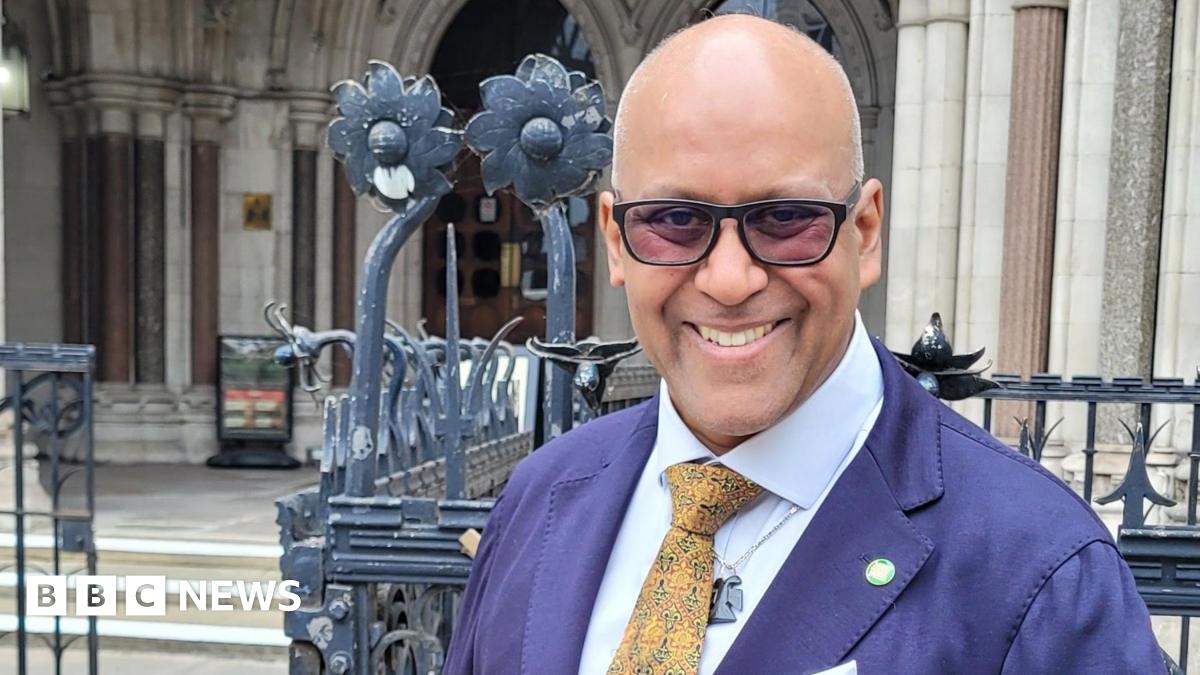 Shahrar Ali stands outside the Royal Courts of Justice, wearing a dark blue blazer, white shirt, and patterned yellow tie. He has glasses and is smiling at the camera. Behind him are ornate iron gates and the stone façade of the historic court building...